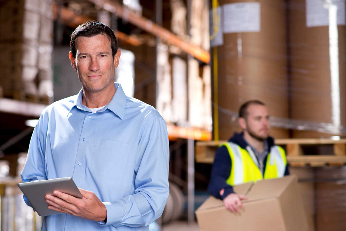 Two men in a warehouse, one with a tablet, the other carrying a cardboard box. 