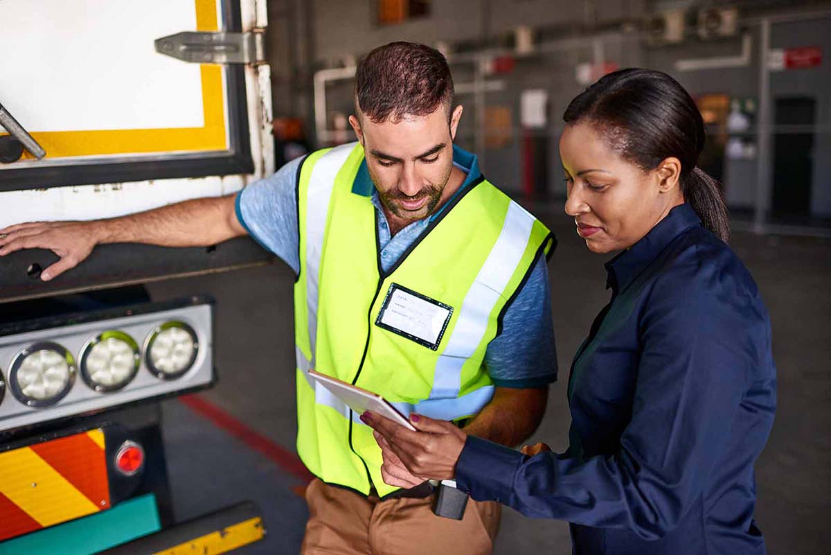 Man in high-viz vest and woman in dark blue standing behind a truck, looking at paperwork.