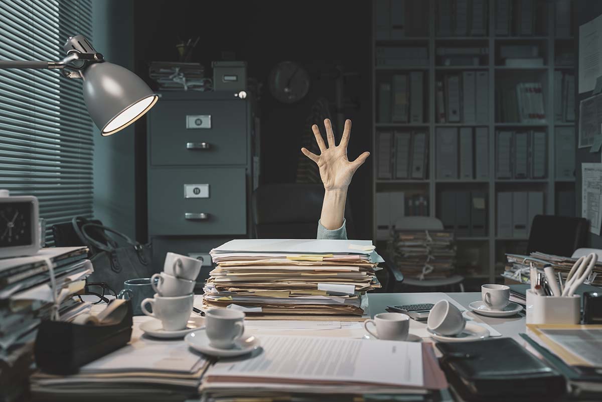 Een bureau vol papierwerk, iemand is daarachter verdwenen en steekt een hand op in nood