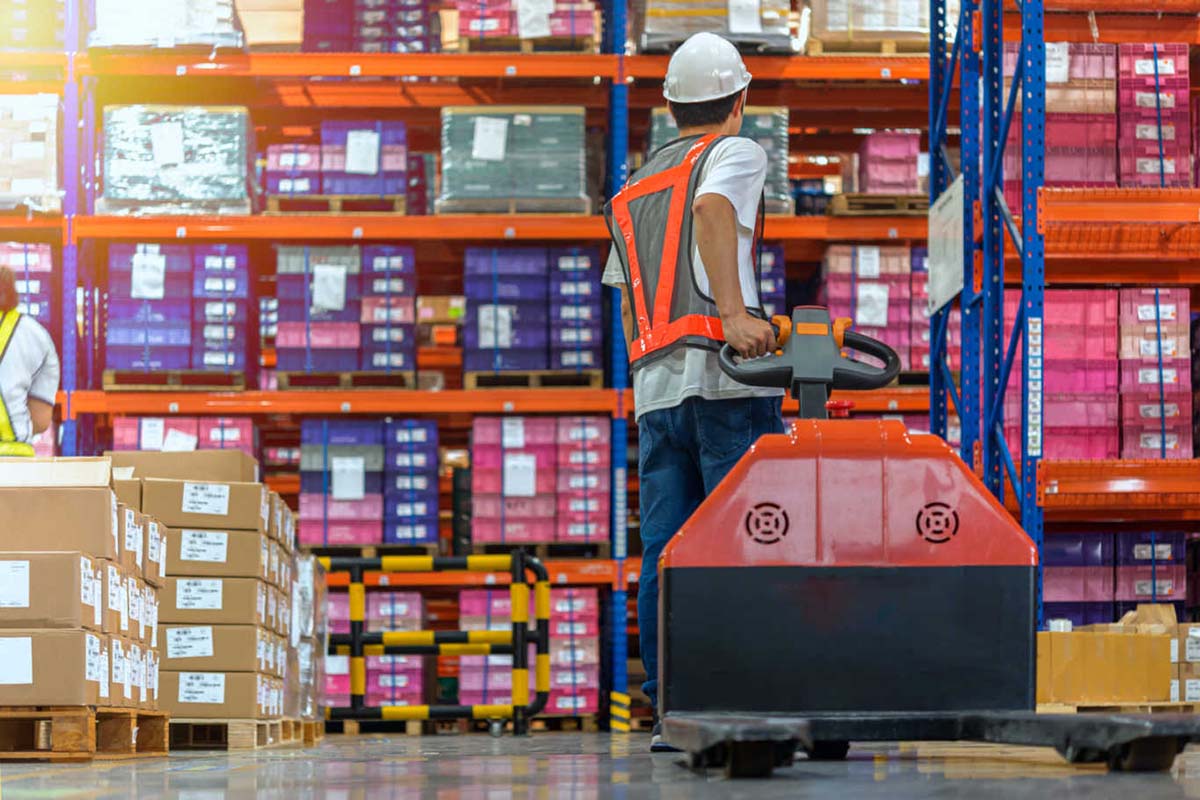 Man in magazijn die een pallettruck achter zich aan trekt. 