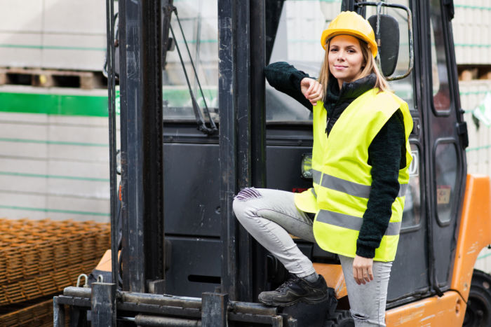Vrouw met helm en lichtgevend hesje bij een heftruck op een bouwplaats